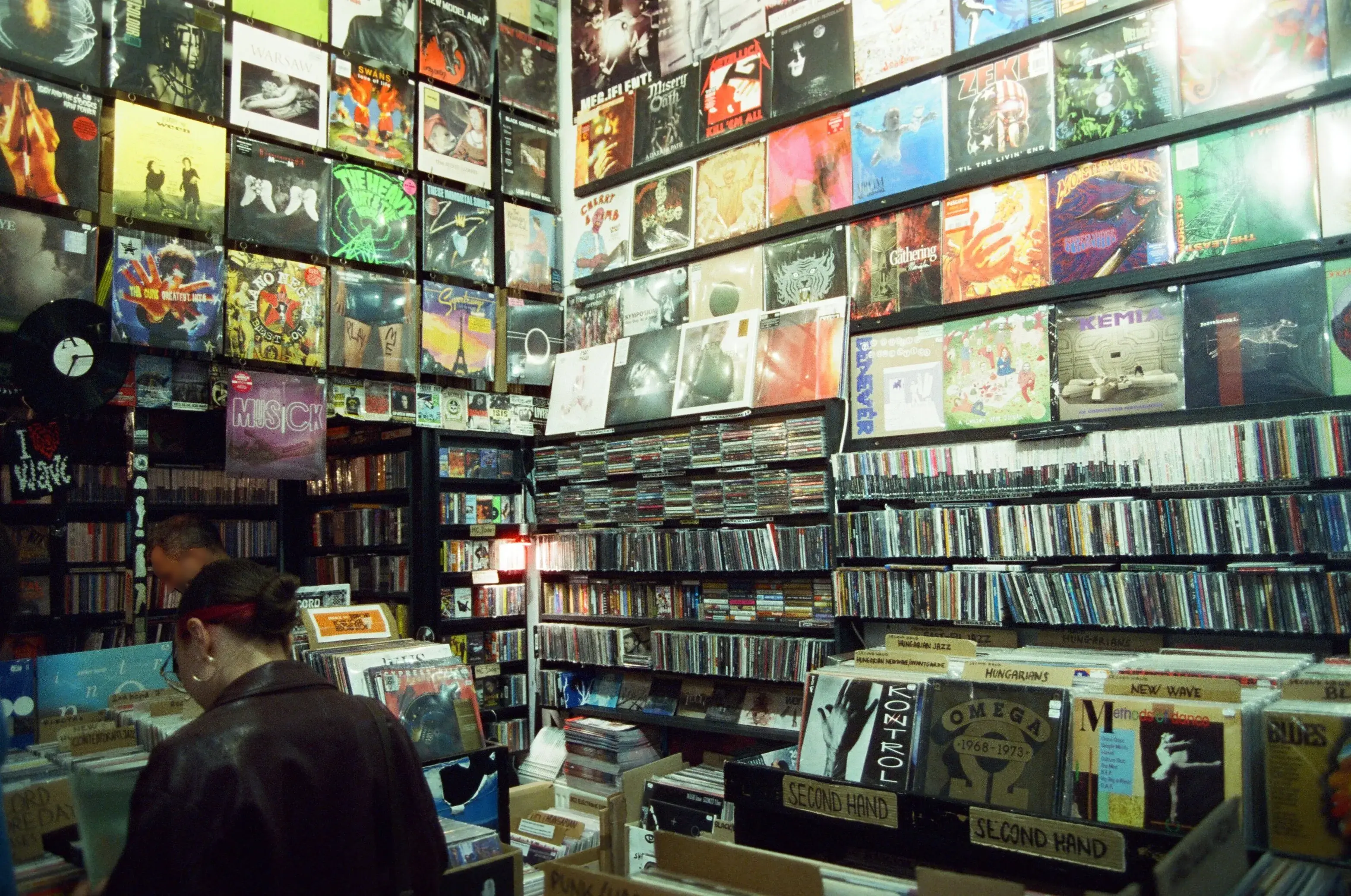 A film photograph inside a Budapest record store: album covers are displayed across tall walls, creating a colorful collage of music artwork. Shelves filled with CDs stretch along the room, while record bins labeled by genre and section fill the floor. A person in a dark jacket and a red streak in their hair browses through vinyl records near the front of the shop.