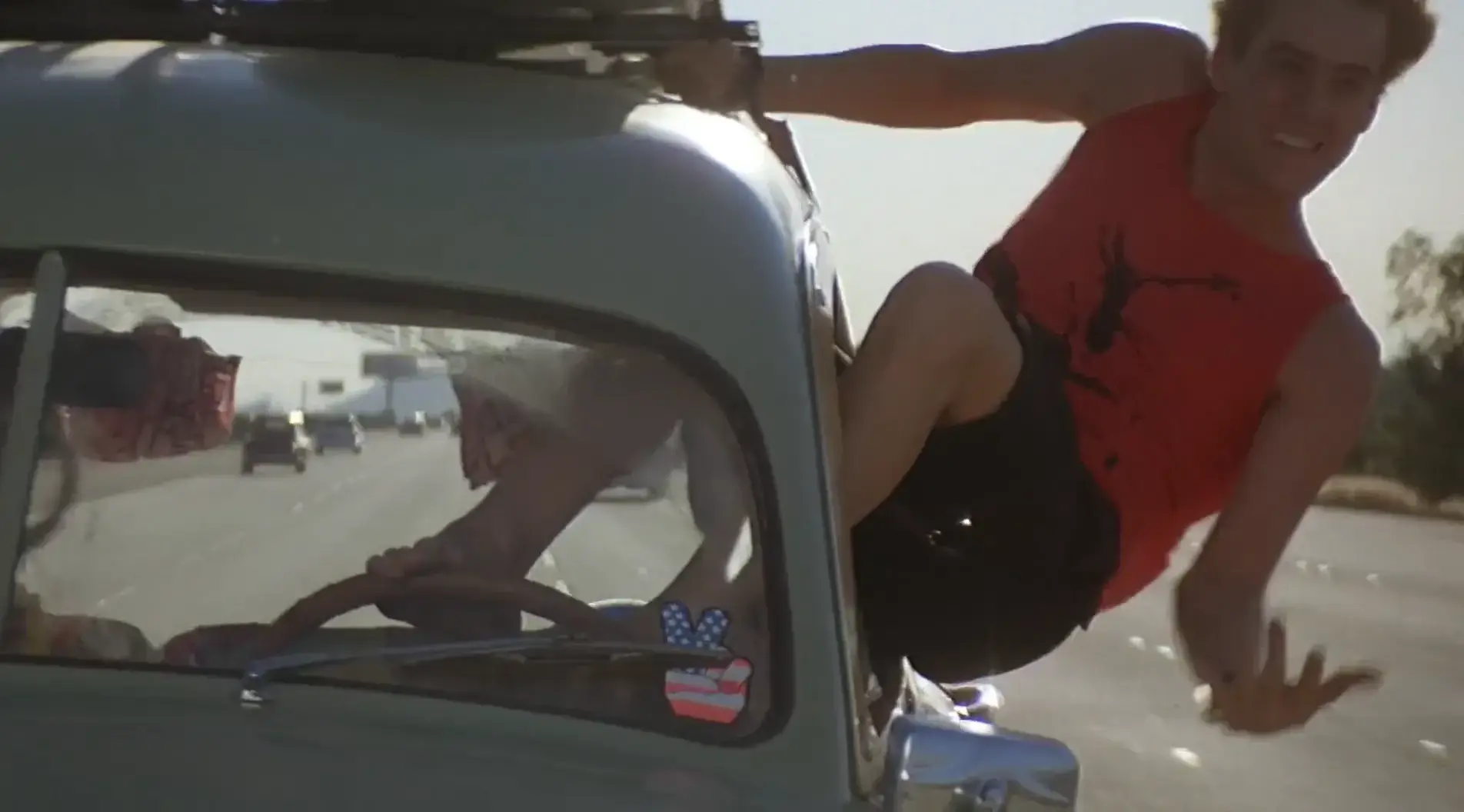 Jim Carrey hanging out of the window of a moving vehicle, his feet controlling the steering wheel